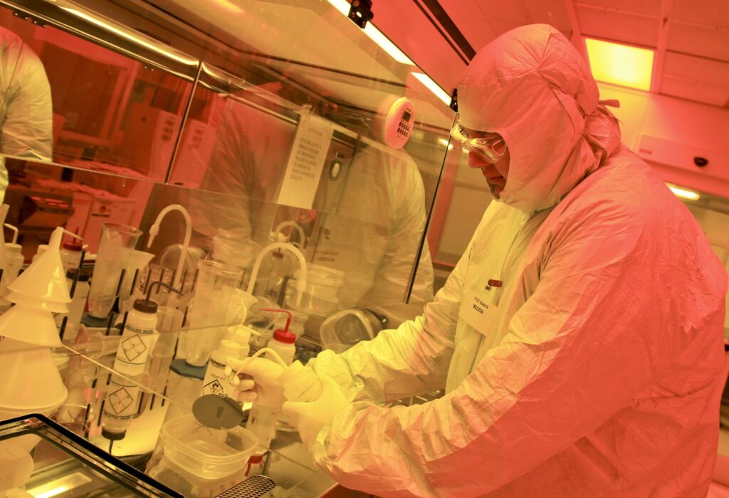 Scientist in a full cleanroom suit rinsing a silicon wafer under yellow light